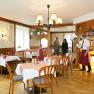 Ein rustikales Restaurant mit Holzm&ouml;beln und Tischdecken. Ein Kellner begr&uuml;&szlig;t eine Familie am Eingang.