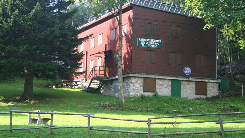 Piros sz&iacute;nű, faburkolatos &eacute;p&uuml;let, f&aacute;kkal &eacute;s r&eacute;ttel k&ouml;r&uuml;lv&eacute;ve, "Naturfreundehaus Ortsgruppe Ternitz" felirattal.