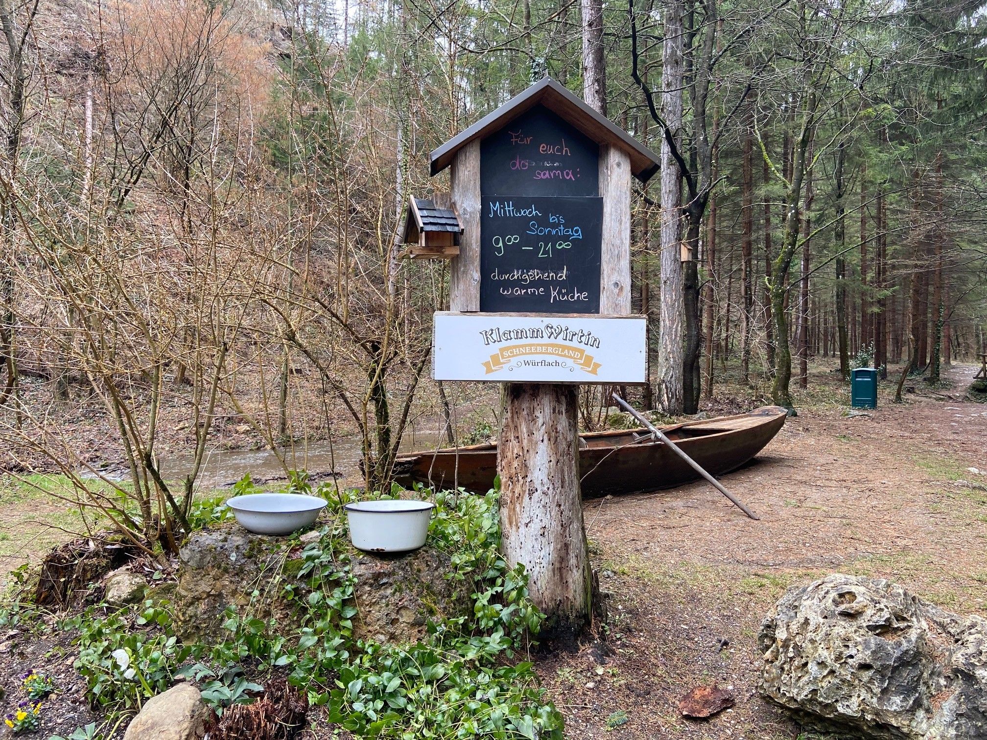 Wegweiser im Wald mit Öffnungszeiten der Klammwirtin, umgeben von Bäumen und einem Boot.