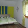 Schlafzimmer mit Doppelbett, gr&uuml;n-gelb gestreifte Wand, gr&uuml;ne Vorh&auml;nge, Holzboden.