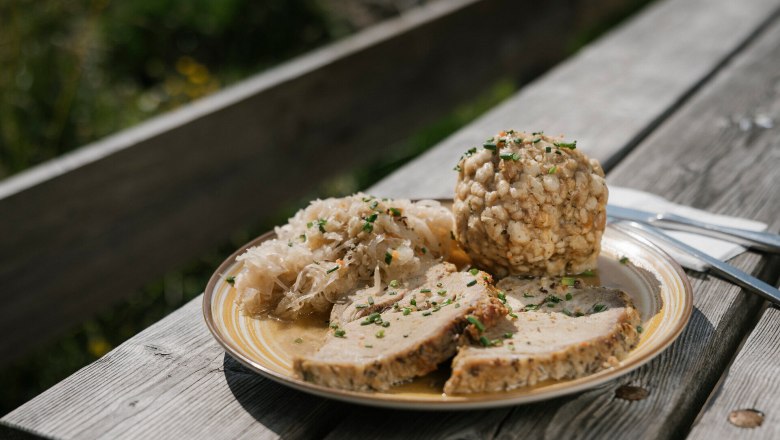 Ein Teller mit Fleischlaibchen, Kn&ouml;del und Sauerkraut auf einem Holztisch im Freien.