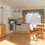 Bright kitchen with dining table and chairs, white cupboards, wooden floor, windows with curtains.