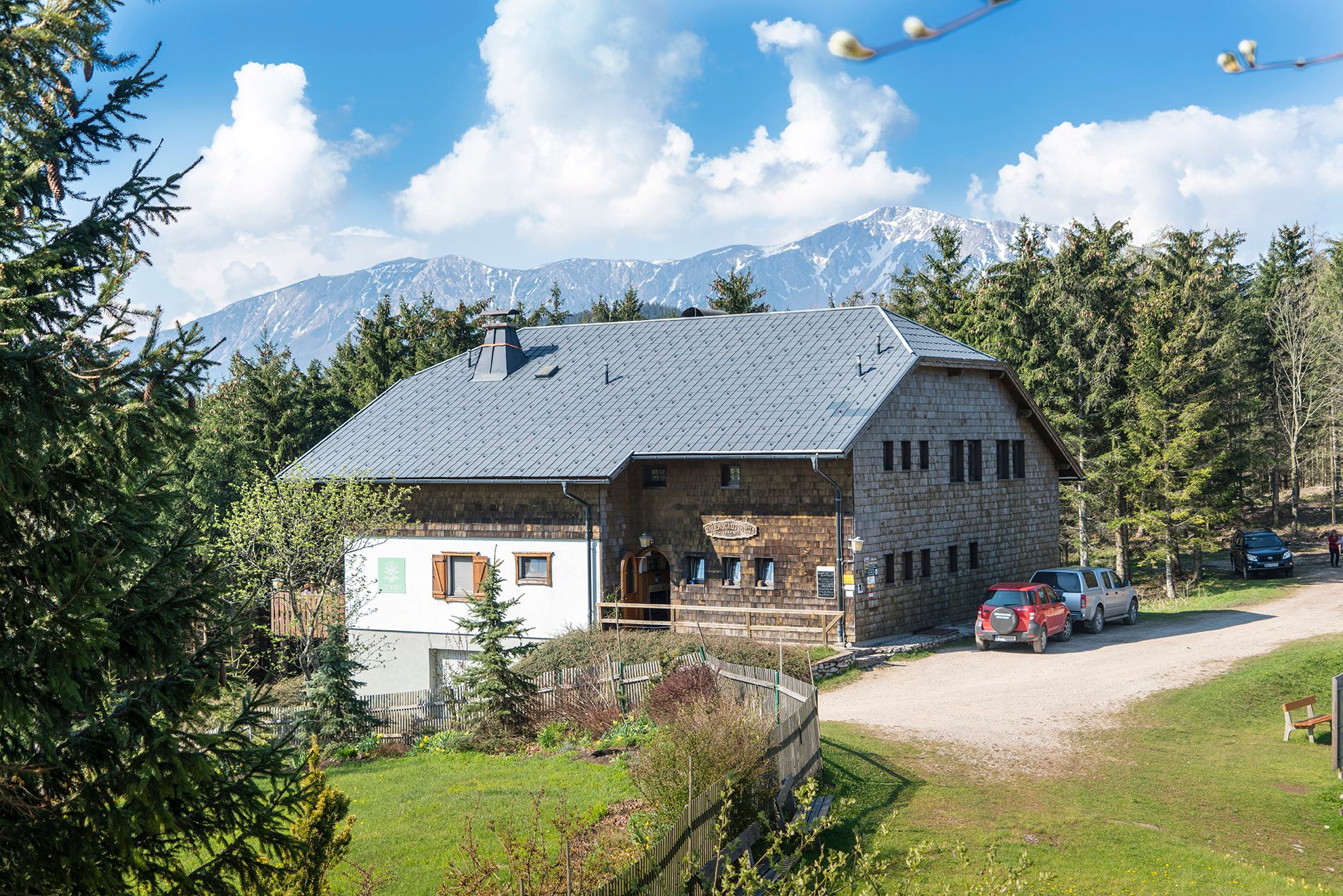Ein rustikales Schutzhaus in einer bergigen Landschaft mit Bäumen und Autos im Vordergrund.