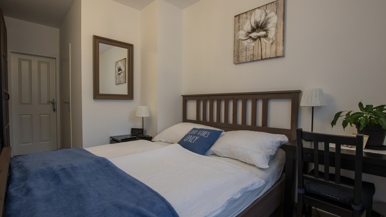 Gem&uuml;tliches Doppelzimmer mit Holzbett, blauer Decke und Dekokissen. Ein Spiegel und ein Blumenbild an der Wand, daneben ein Schreibtisch mit Pflanze.