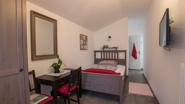 Ein gem&uuml;tliches Einzelzimmer mit Holzm&ouml;beln, einem Bett, einem kleinen Tisch mit St&uuml;hlen, einem Spiegel und einem Fernseher an der Wand.