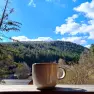 Eine Tasse steht auf einem Gel&auml;nder mit Blick auf bewaldete H&uuml;gel und blauen Himmel.