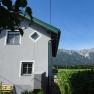 Haus mit grauer Fassade und gr&uuml;nen Fensterrahmen, im Hintergrund Berge und blauer Himmel.