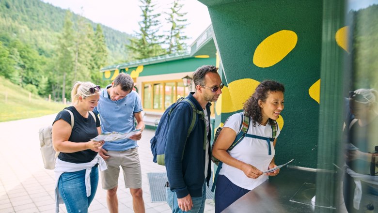 Vier Personen stehen an einem Ticketschalter im Freien, umgeben von grüner Landschaft.