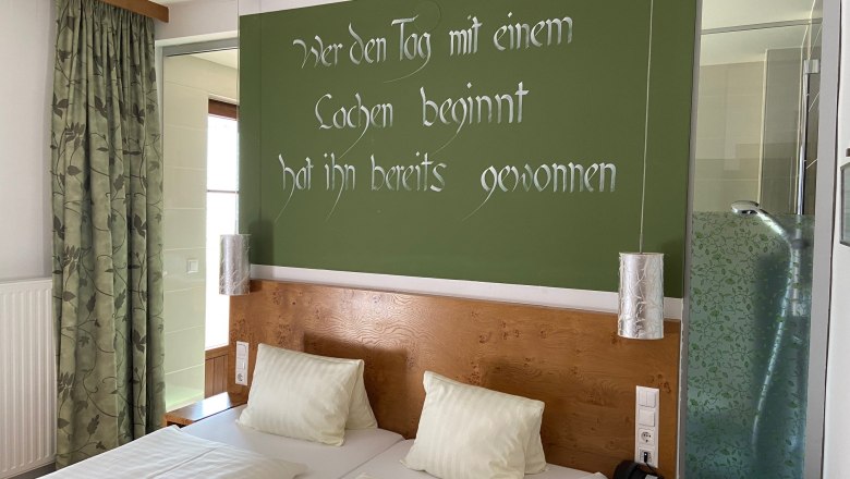 Ein Doppelzimmer mit zwei Betten, grüner Wand mit Spruch, Vorhängen und einer Dusche mit Glaswand.