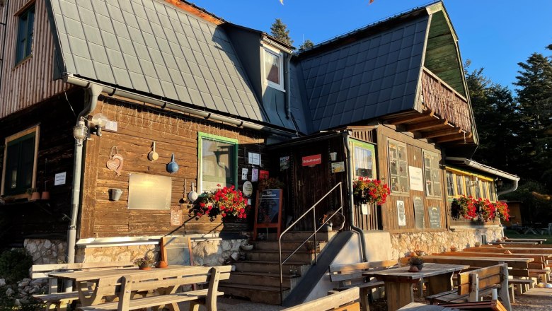 A rustic wooden house with flowers and benches outside in the sunshine.