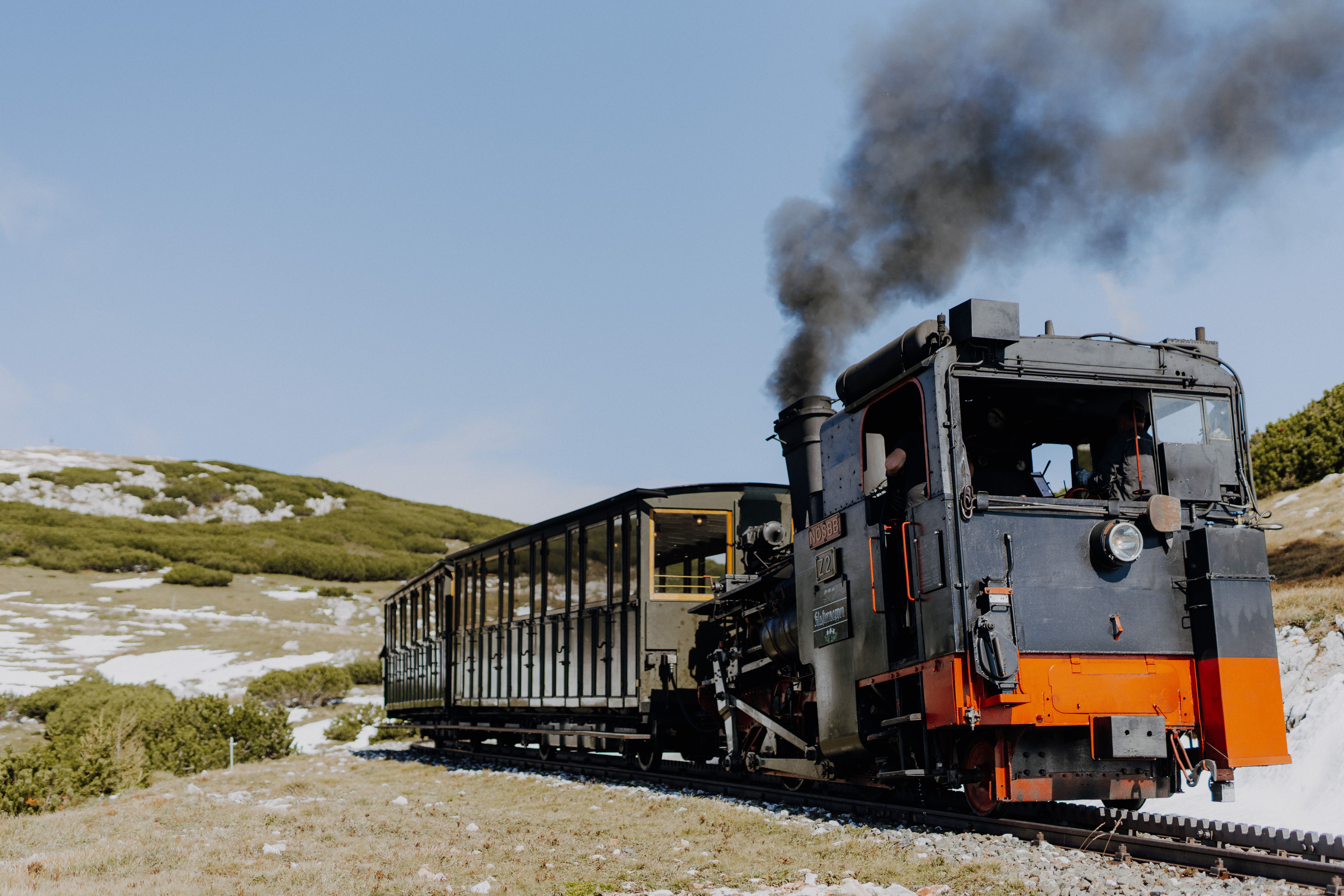 Historische Dampflokomotive mit schwarzem Rauch zieht einen Personenwagen durch eine alpine Landschaft mit Wiesen, Felsen und letzten Schneeresten.