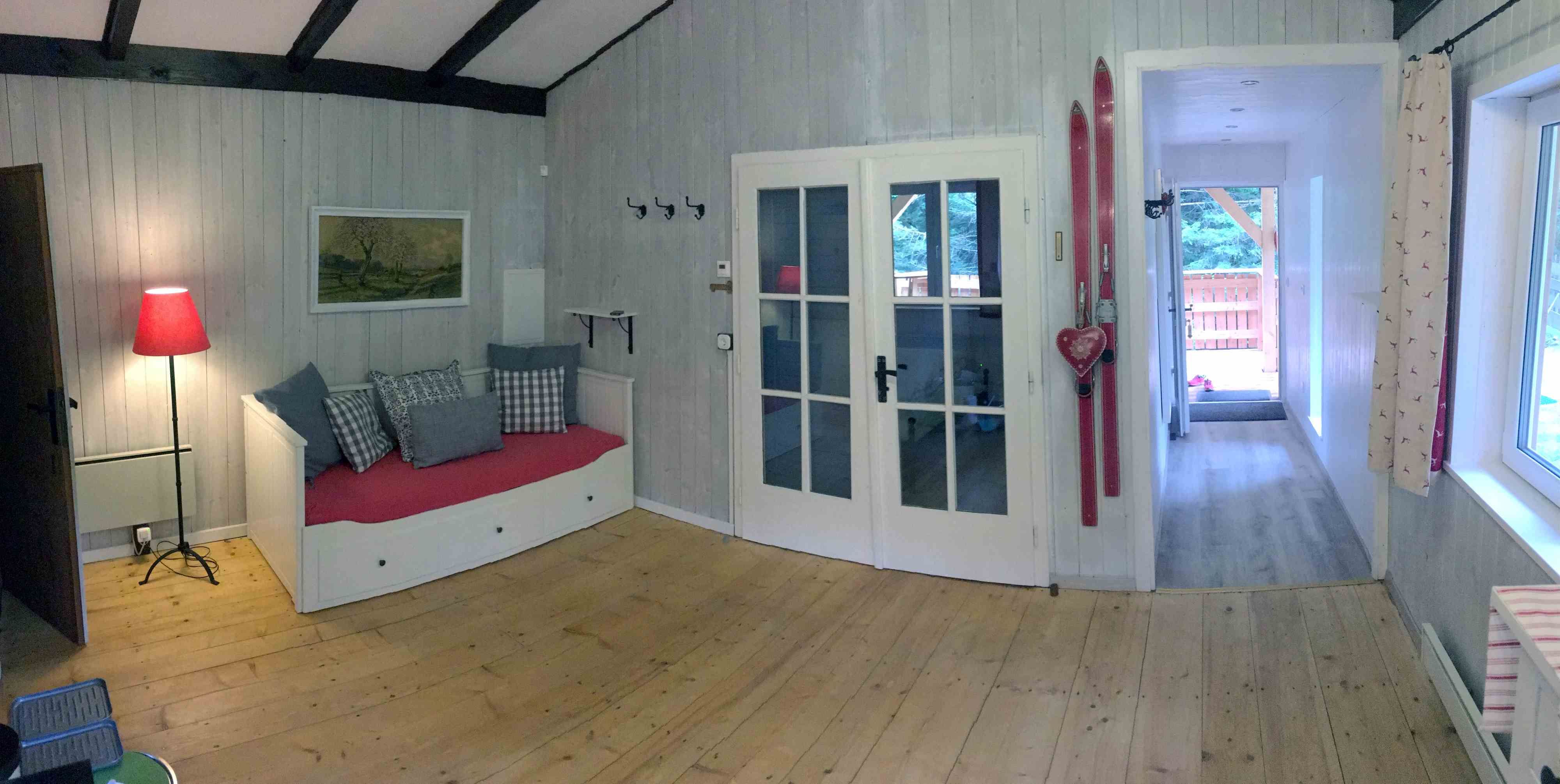 Innenansicht eines gemütlichen Ferienhauses mit Holzboden, rotem Sofa, Skiern an der Wand und Glastüren.