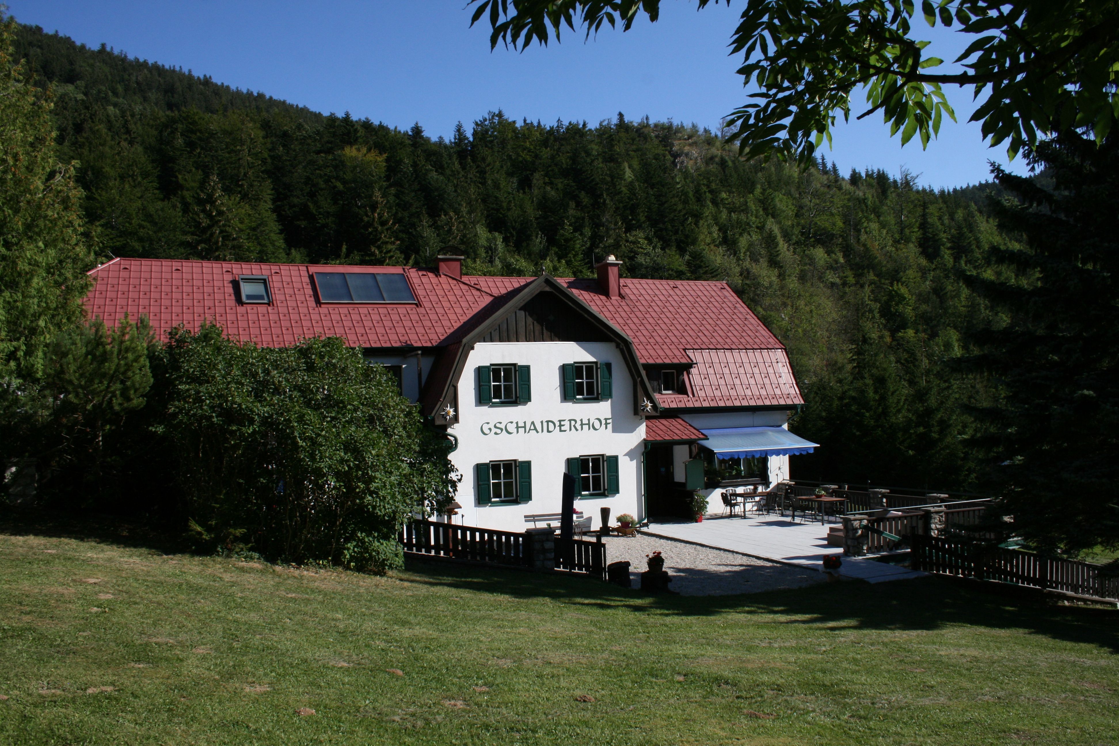 Ein traditionelles Haus mit rotem Dach, umgeben von Wald und Wiese, mit der Aufschrift 'Gschaiderhof'.