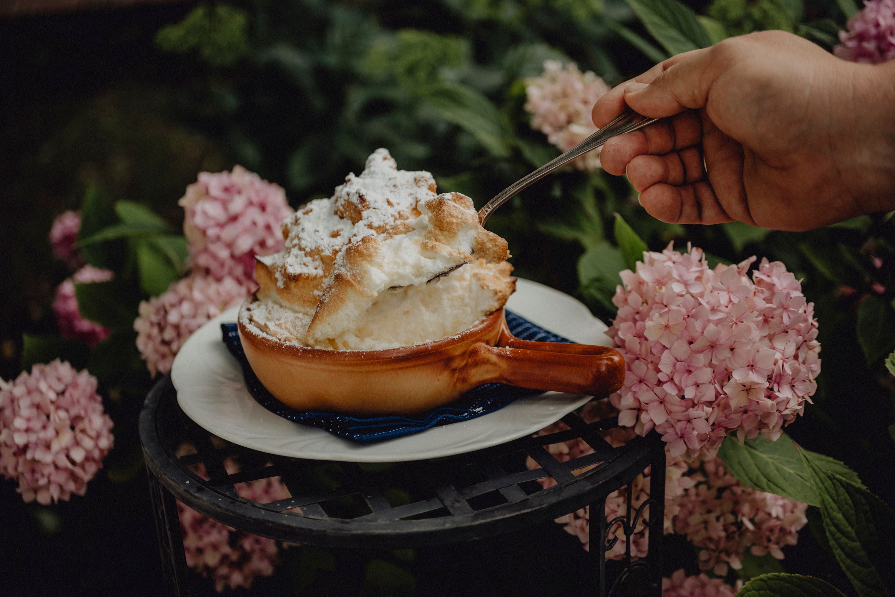 Ein Soufflé in einer braunen Schale, umgeben von rosa Hortensien, wird mit einem Löffel serviert.
