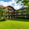 Ein traditionelles Hotel mit Holzbalkonen und einem gepflegten Garten im Vordergrund, umgeben von B&auml;umen und Bergen im Hintergrund.