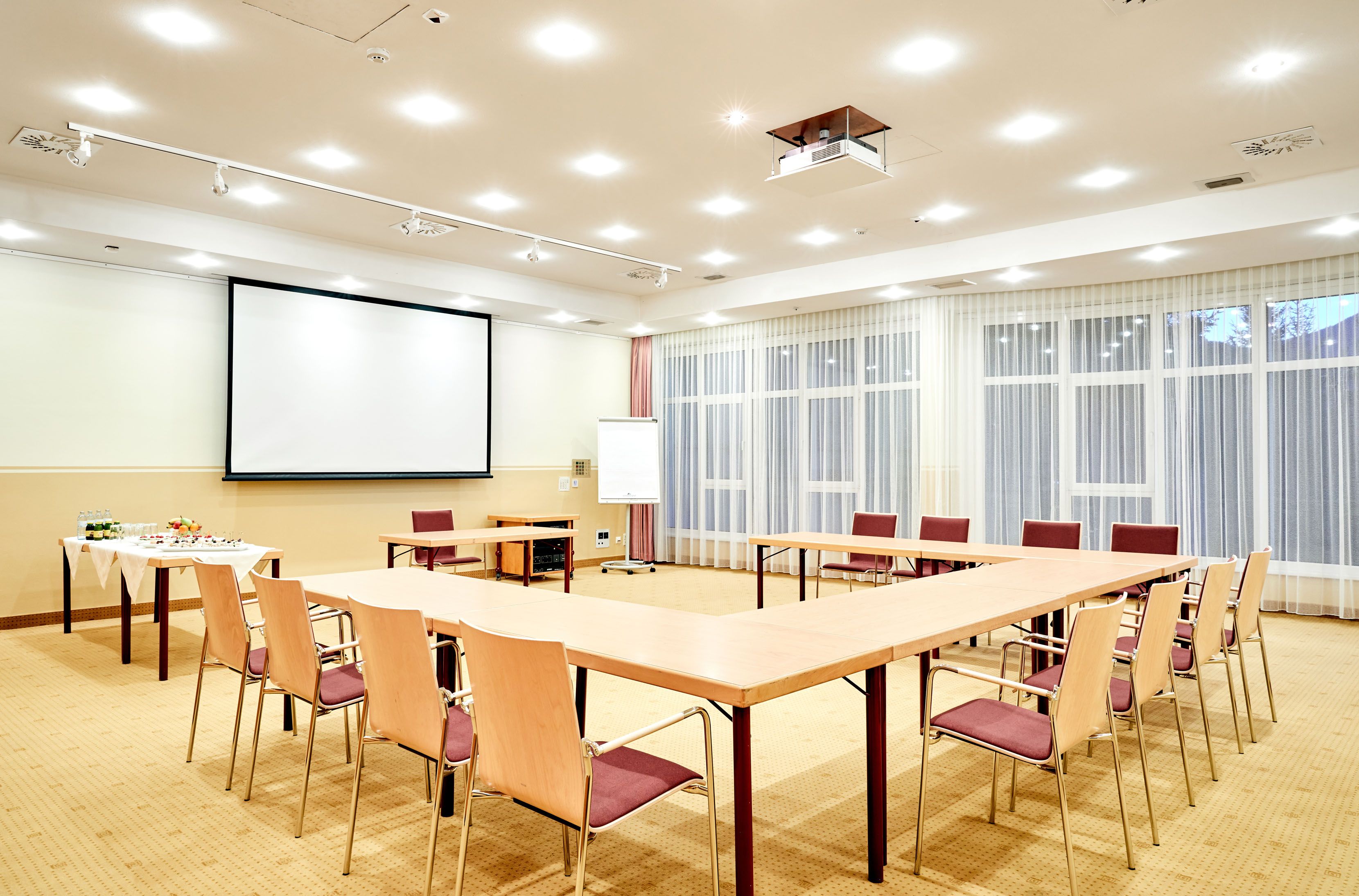 Ein heller Seminarraum mit U-förmiger Tischanordnung, Leinwand, Flipchart und Getränketisch.