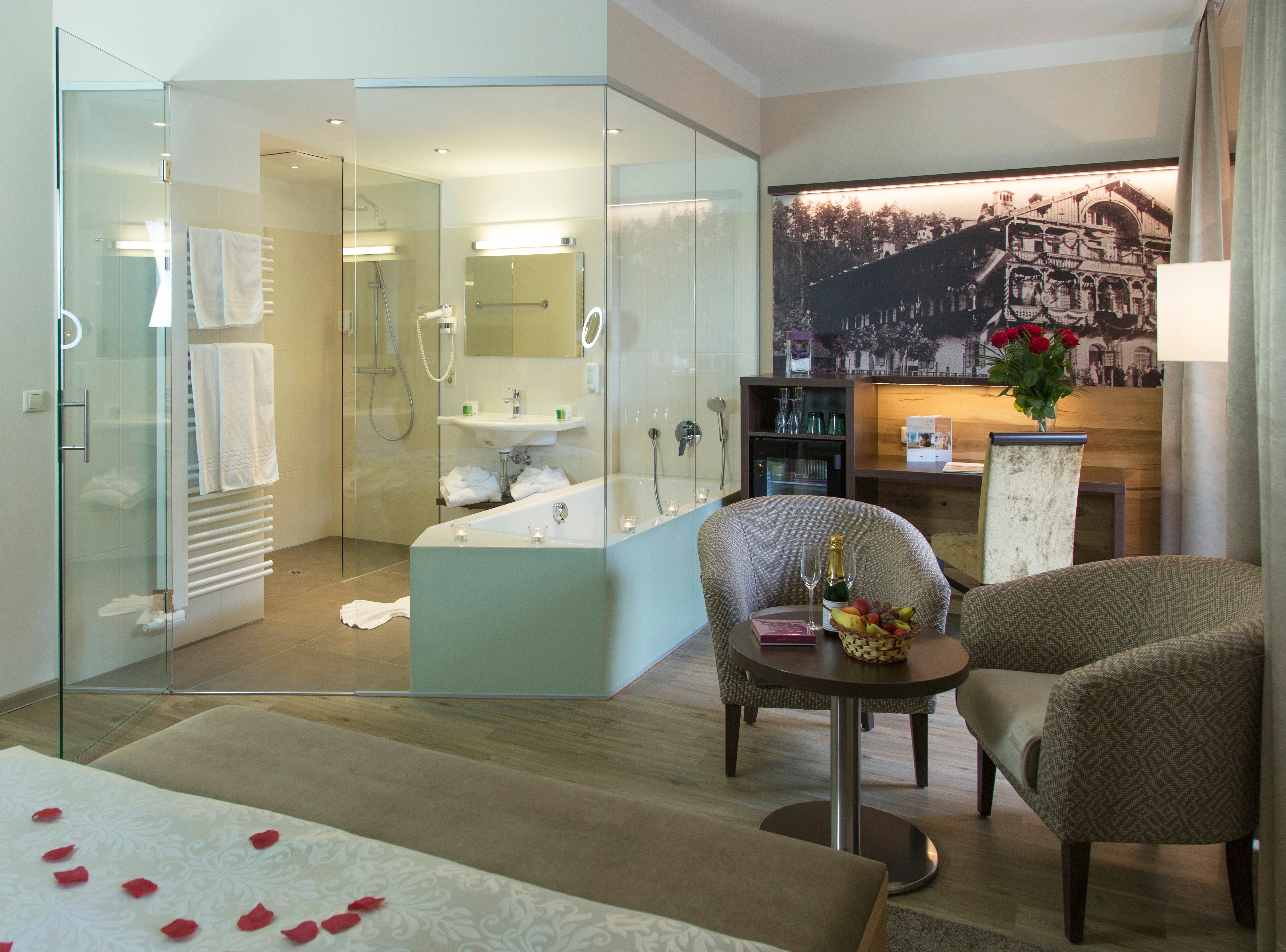 Luxuriöses Hotelzimmer mit Glasbad, gemütlichen Sesseln, Tisch mit Champagner und Früchten, sowie einem großen Bett mit Rosenblättern.