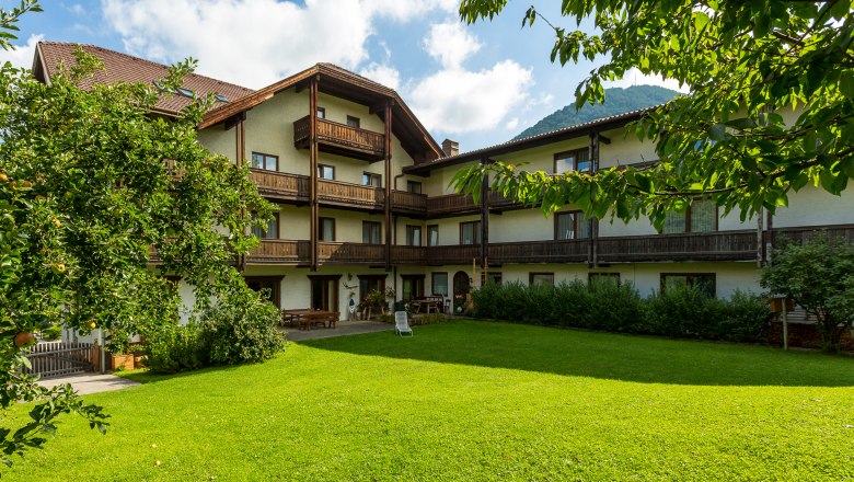 A traditional hotel with wooden balconies and a well-tended garden in the foreground, surrounded by trees and mountains in the background.