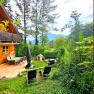 A vacation home with a terrace and garden furniture, surrounded by trees and green vegetation. Mountains can be seen in the background.