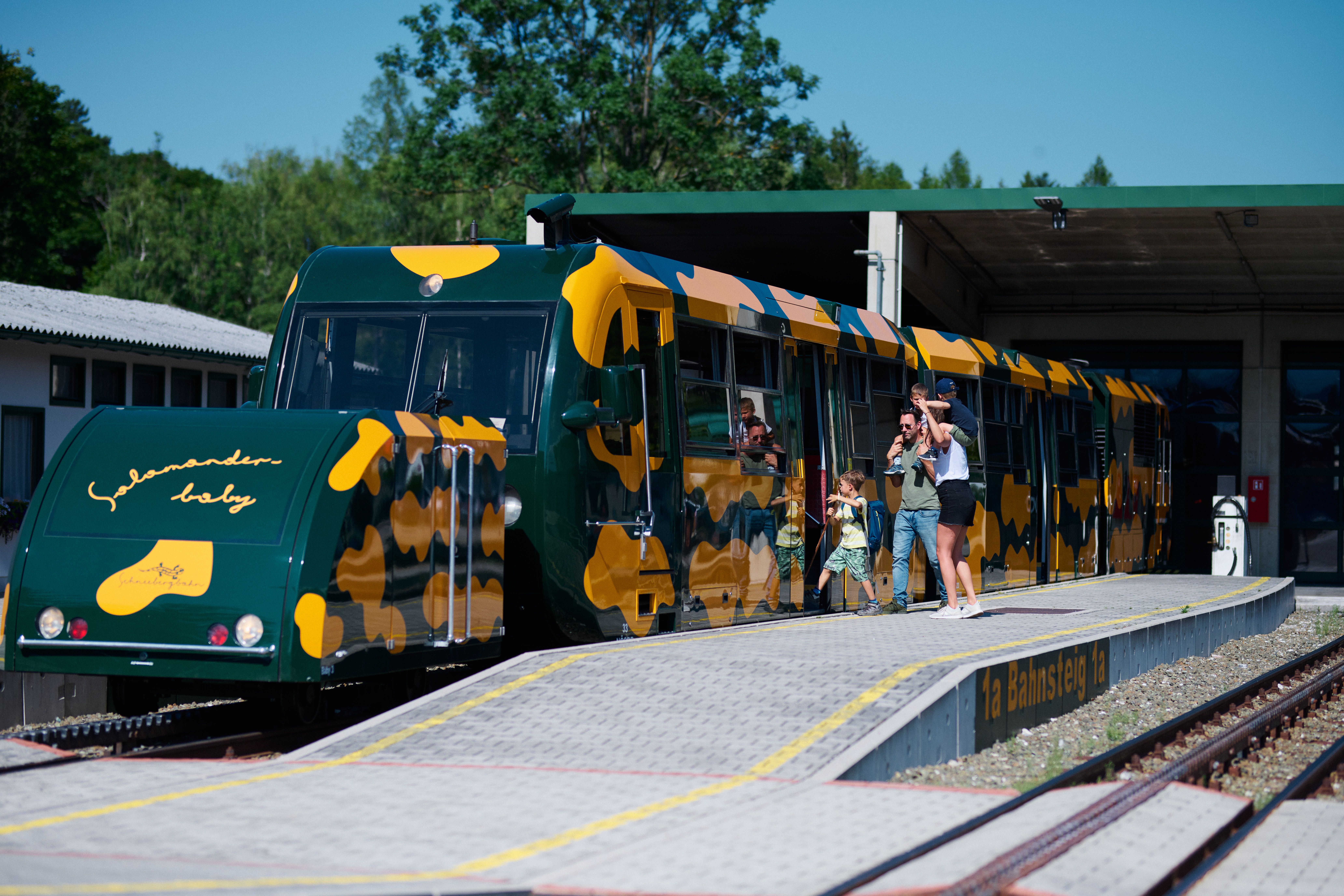Gelb-grüner Salamanderzug steht an einem Bahnsteig, während Fahrgäste ein- und aussteigen. Im Hintergrund sind ein Bahnhofsgebäude und Bäume zu sehen.
