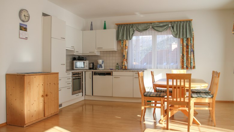 Bright kitchen with dining table and chairs, white cupboards, wooden floor, windows with curtains.