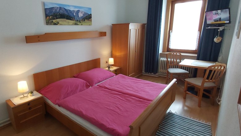 A cozy double room with wooden furniture, a double bed with pink bed linen, a table with two chairs and a small TV.