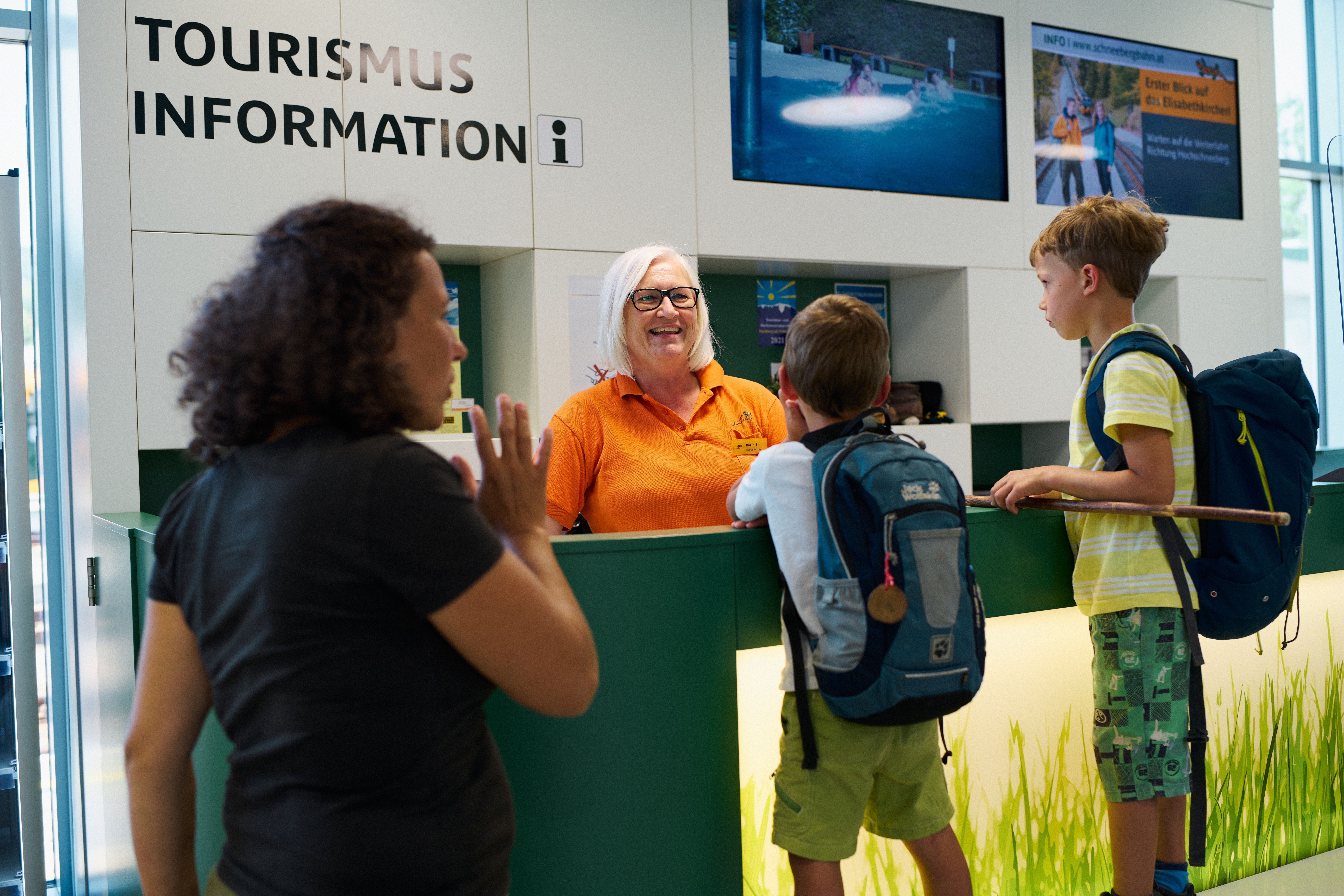 Zwei Jungen mit Rucksäcken im Gespräch mit einer Mitarbeiterin an der Tourismusinformation.