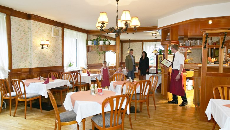 A rustic restaurant with wooden furniture and tablecloths. A waiter greets a family at the entrance.