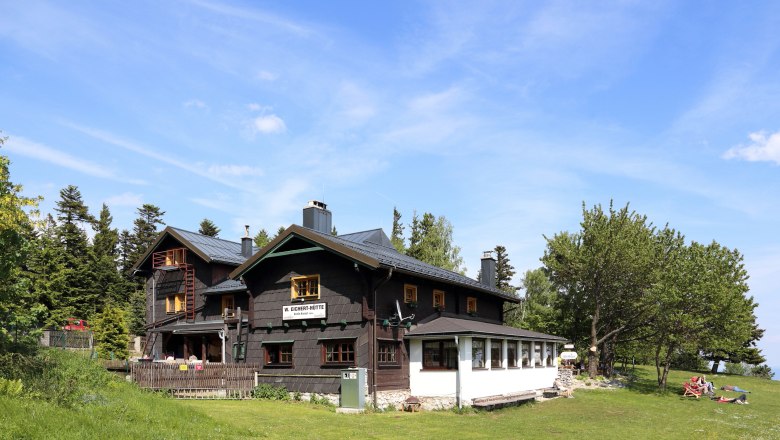 A mountain hut with dark wood and white windows, surrounded by trees and meadows, under a blue sky.