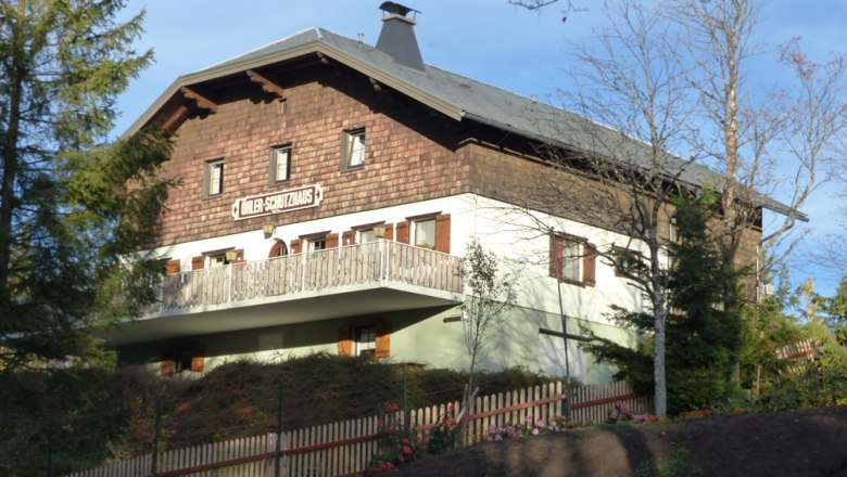 Ein traditionelles Geb&auml;ude mit Holzverkleidung und Balkon, umgeben von B&auml;umen und einem Holzzaun. Schild: "D&ouml;hler-Schutzhaus".