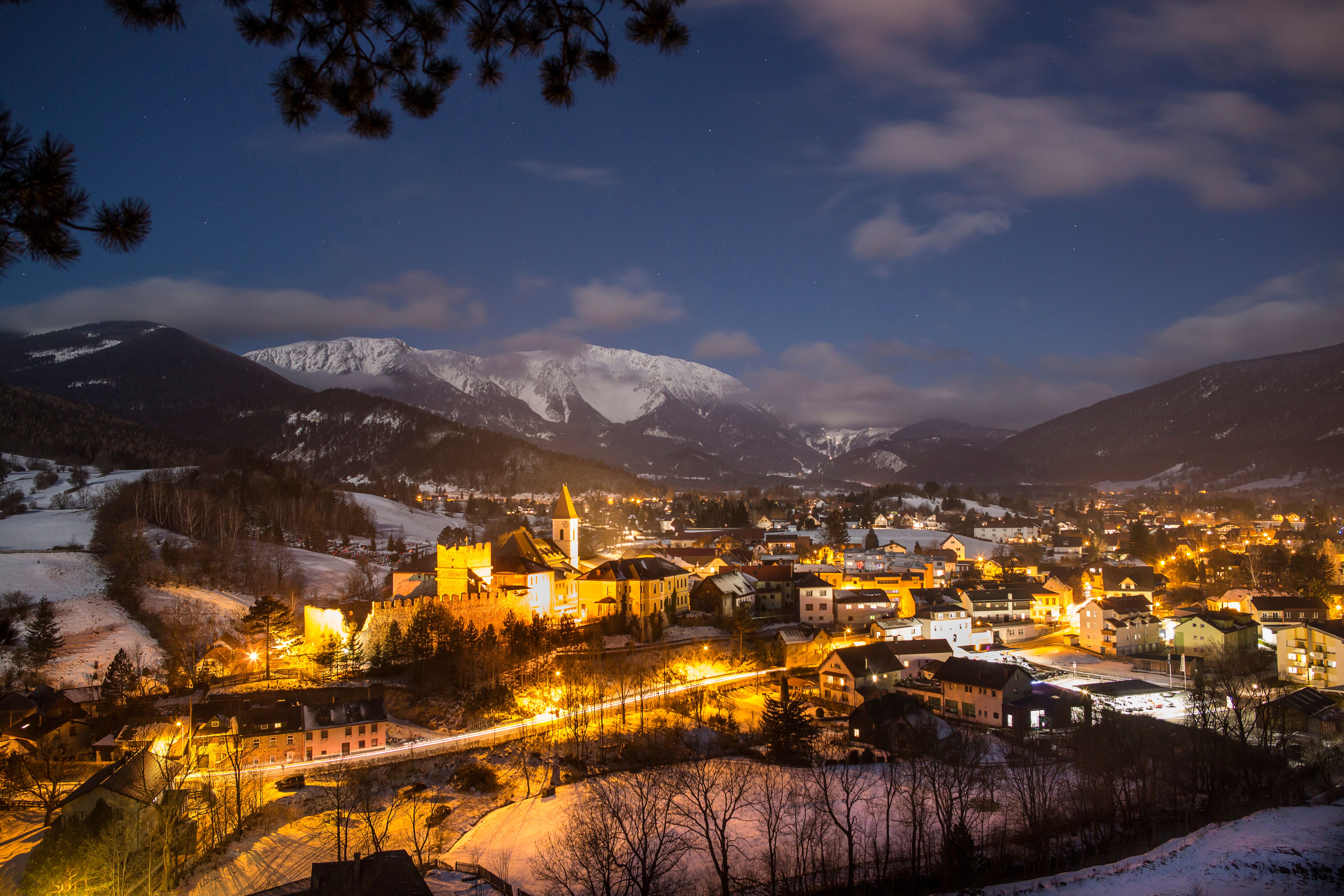 Winterliche Nachtaufnahme einer beleuchteten Bergstadt mit schneebedeckten Hügeln und Bergen im Hintergrund