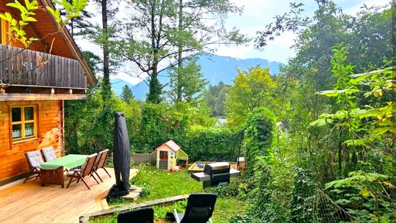 A vacation home with a terrace and garden furniture, surrounded by trees and green vegetation. Mountains can be seen in the background.