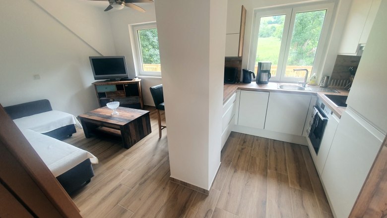 Modern eat-in kitchen with wooden floor, sofa, TV, coffee table and white fitted kitchen with window.