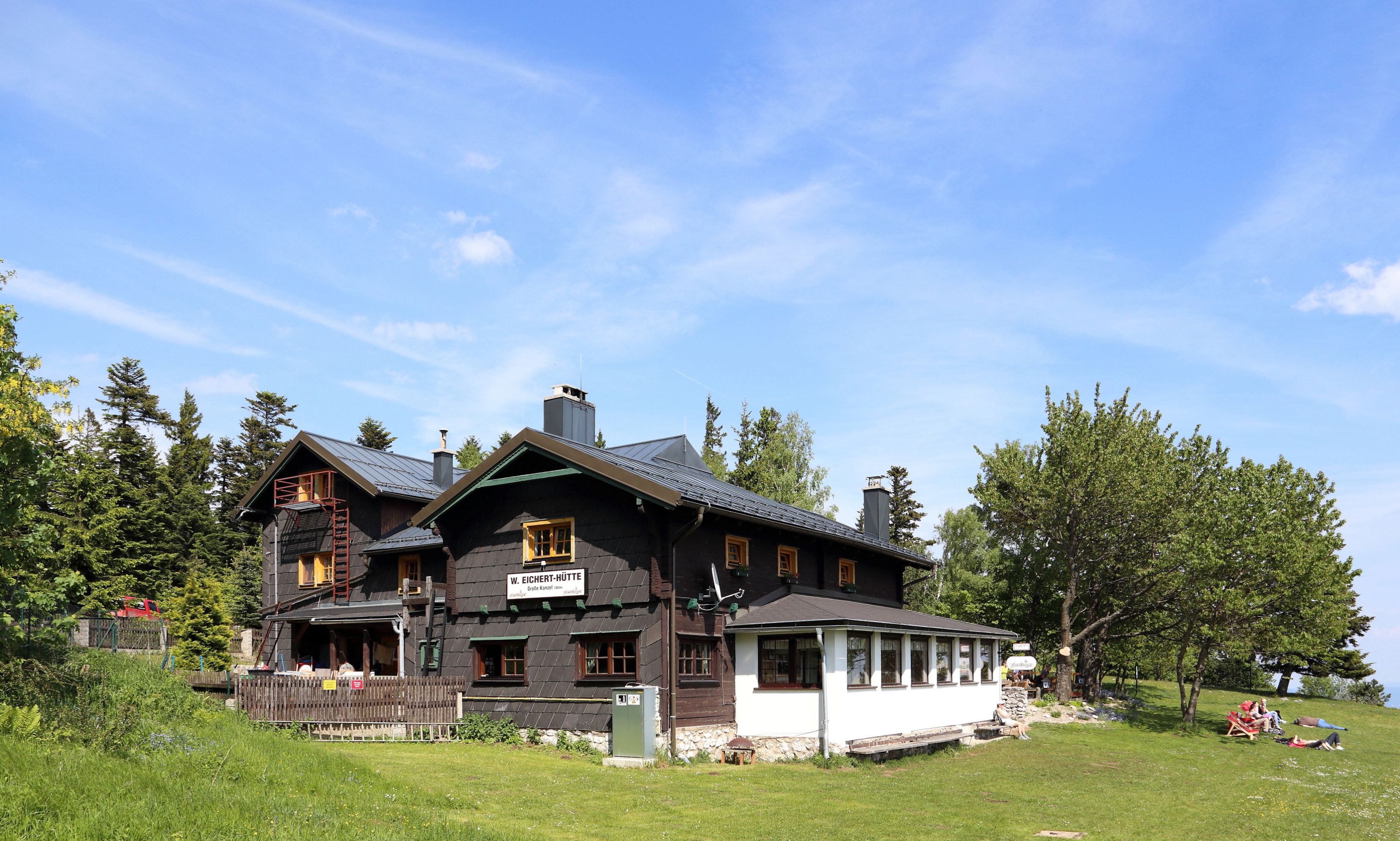 Eine Berghütte mit dunklem Holz und weißen Fenstern, umgeben von Bäumen und Wiese, unter blauem Himmel.