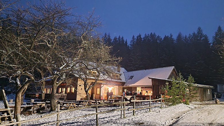 Beleuchtetes Gasthaus mit schneebedecktem Dach und Boden, umgeben von Bäumen und Tischen im Freien bei Abenddämmerung