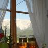 Blick aus einem Fenster mit Vorh&auml;ngen auf einen Berg. Zwei Gl&auml;ser mit Fl&uuml;ssigkeit stehen auf der Fensterbank.