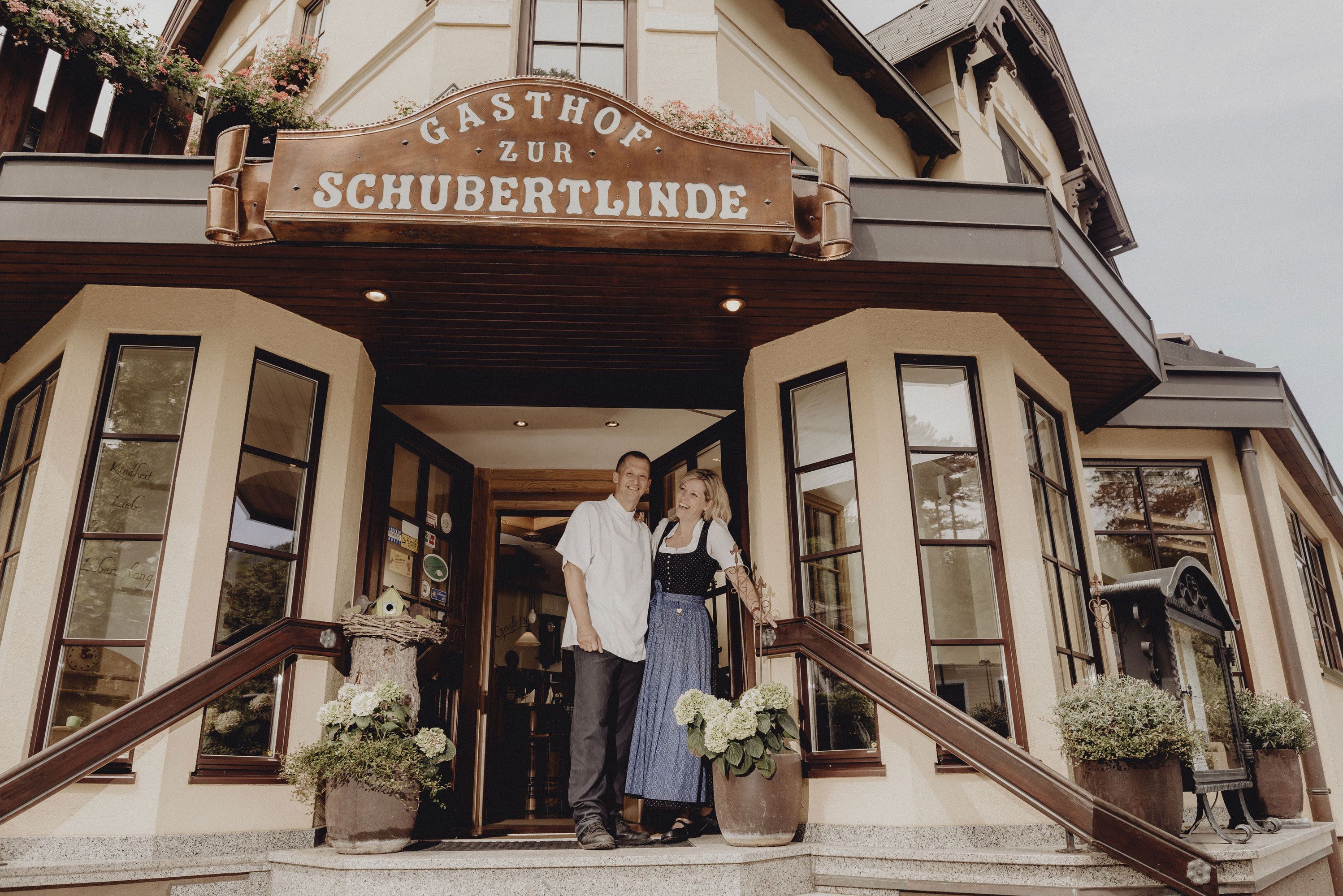 Eingang des Gasthofs Zur Schubertlinde mit zwei Personen in traditioneller Kleidung.