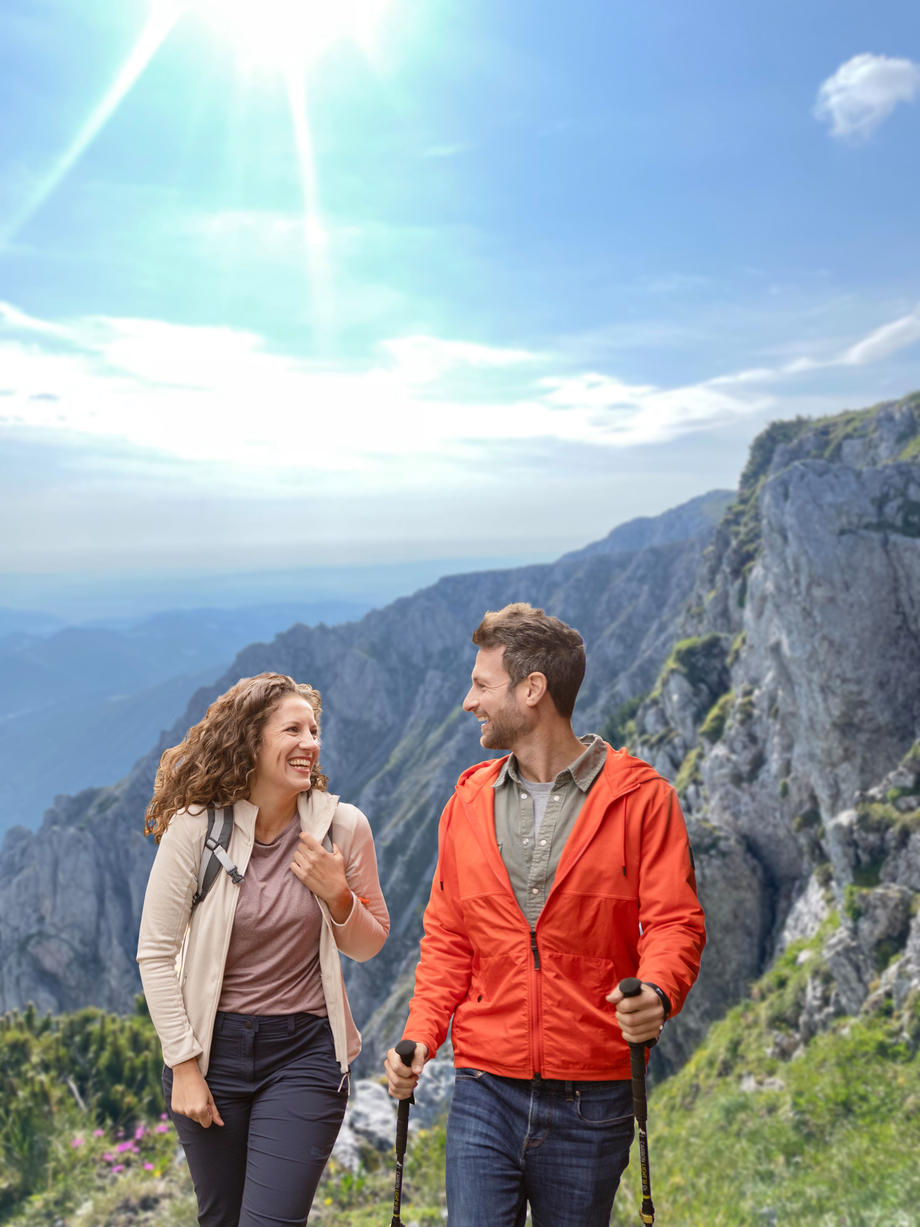 Ein lächelndes Paar wandert bei strahlendem Sonnenschein in den Bergen, schaut sich gut gelaunt an und trägt Outdoor-Kleidung sowie Wanderstöcke vor einer imposanten Felskulisse.
